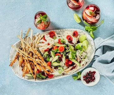 Plate of cranberry-chilli prawns with fresh salad, lime slices, and refreshing drinks on a light blue table.