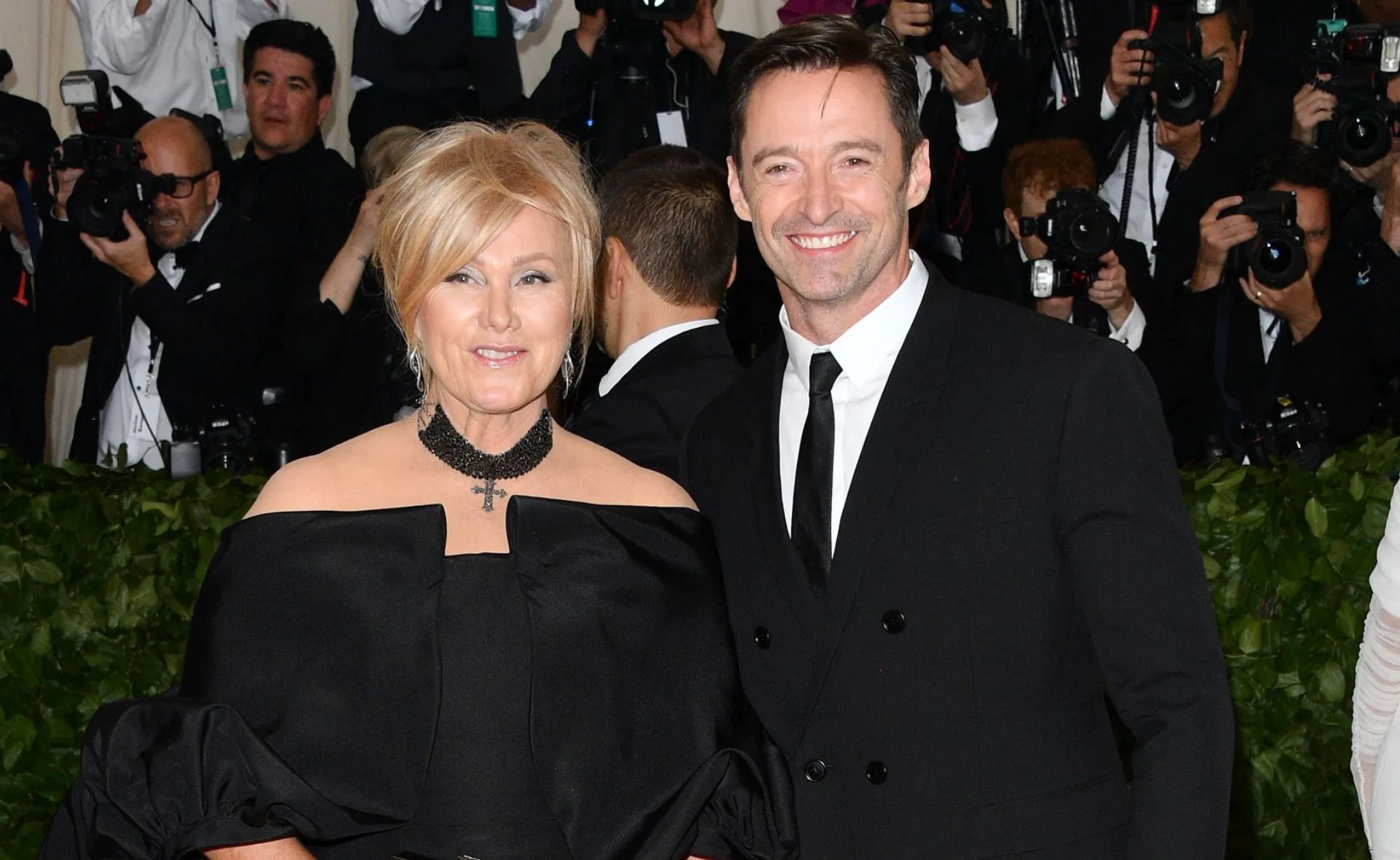 Man and woman in formal attire smiling at an event, surrounded by photographers.