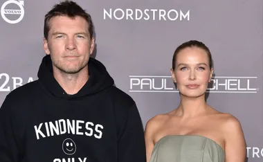 Man in black hoodie with "Kindness Only" text and woman in grey strapless dress at an event with branded backdrop.