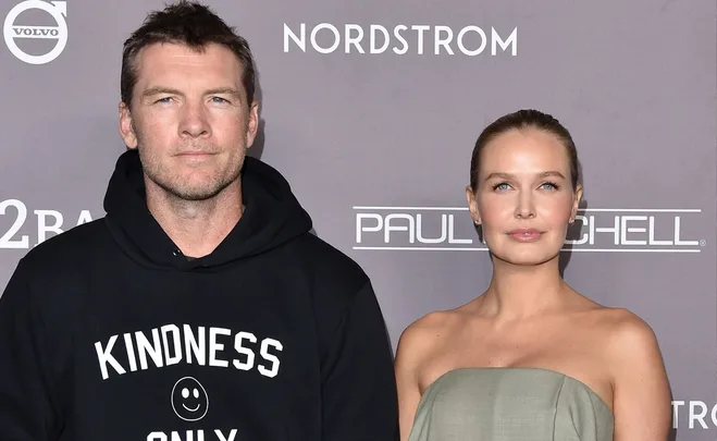 Man in black hoodie with "Kindness Only" text and woman in grey strapless dress at an event with branded backdrop.