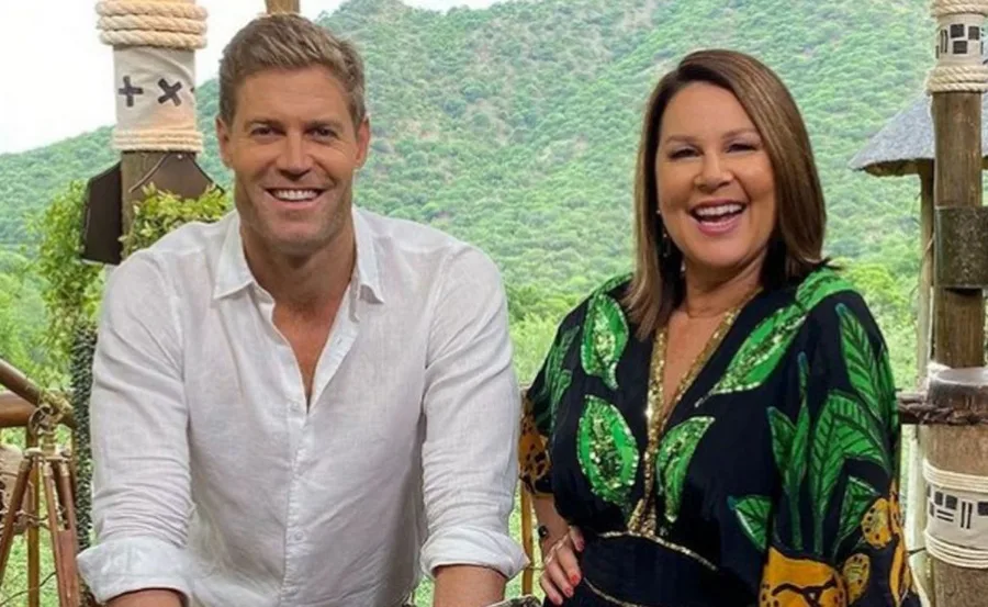 Two hosts smiling on set with a scenic, green landscape in the background.