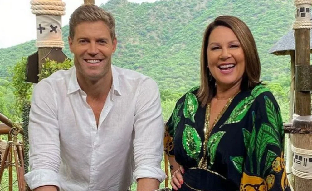 Two hosts smiling on set with a scenic, green landscape in the background.