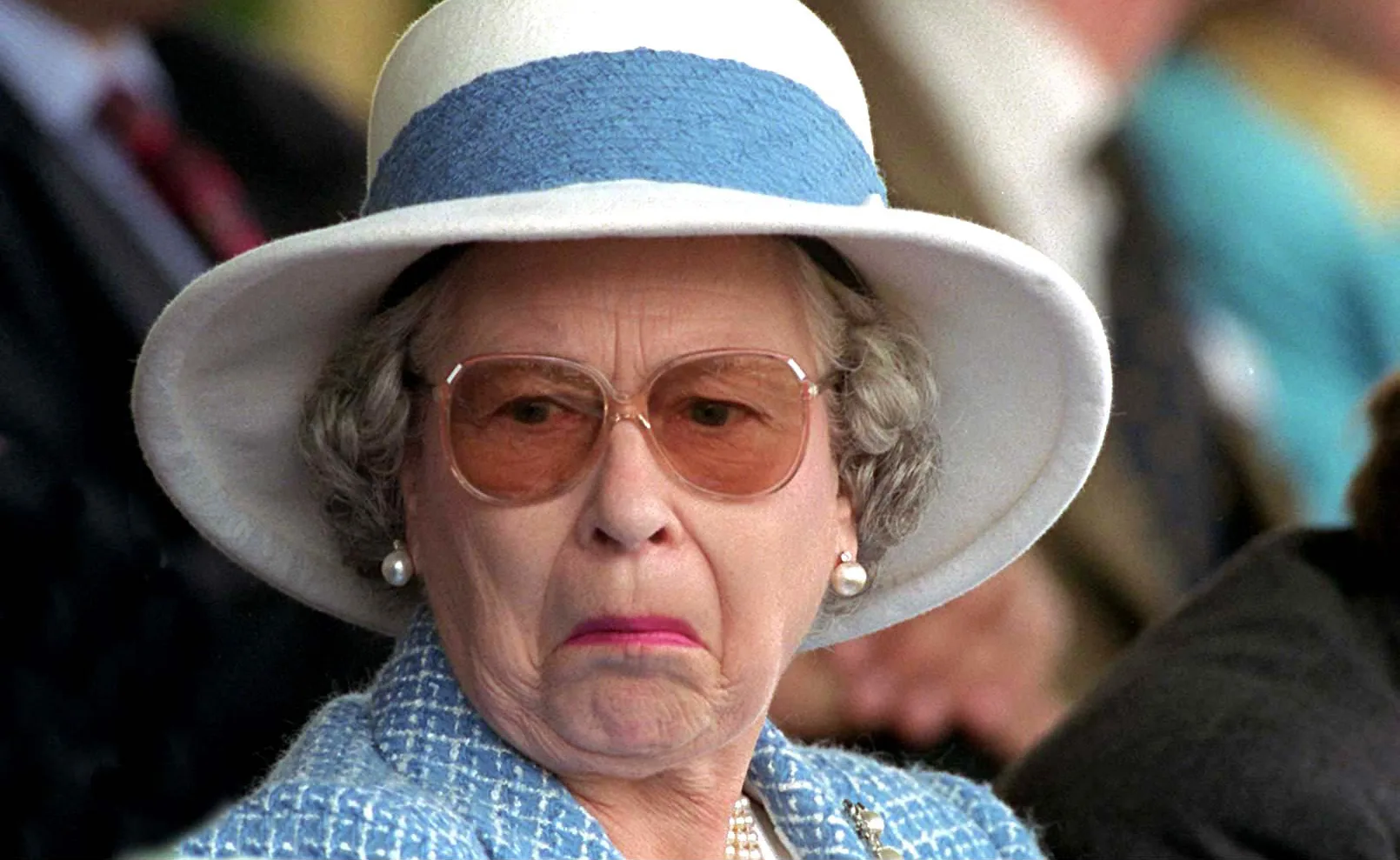 Woman in a blue outfit and white hat making a facial expression, wearing glasses and pearl earrings.