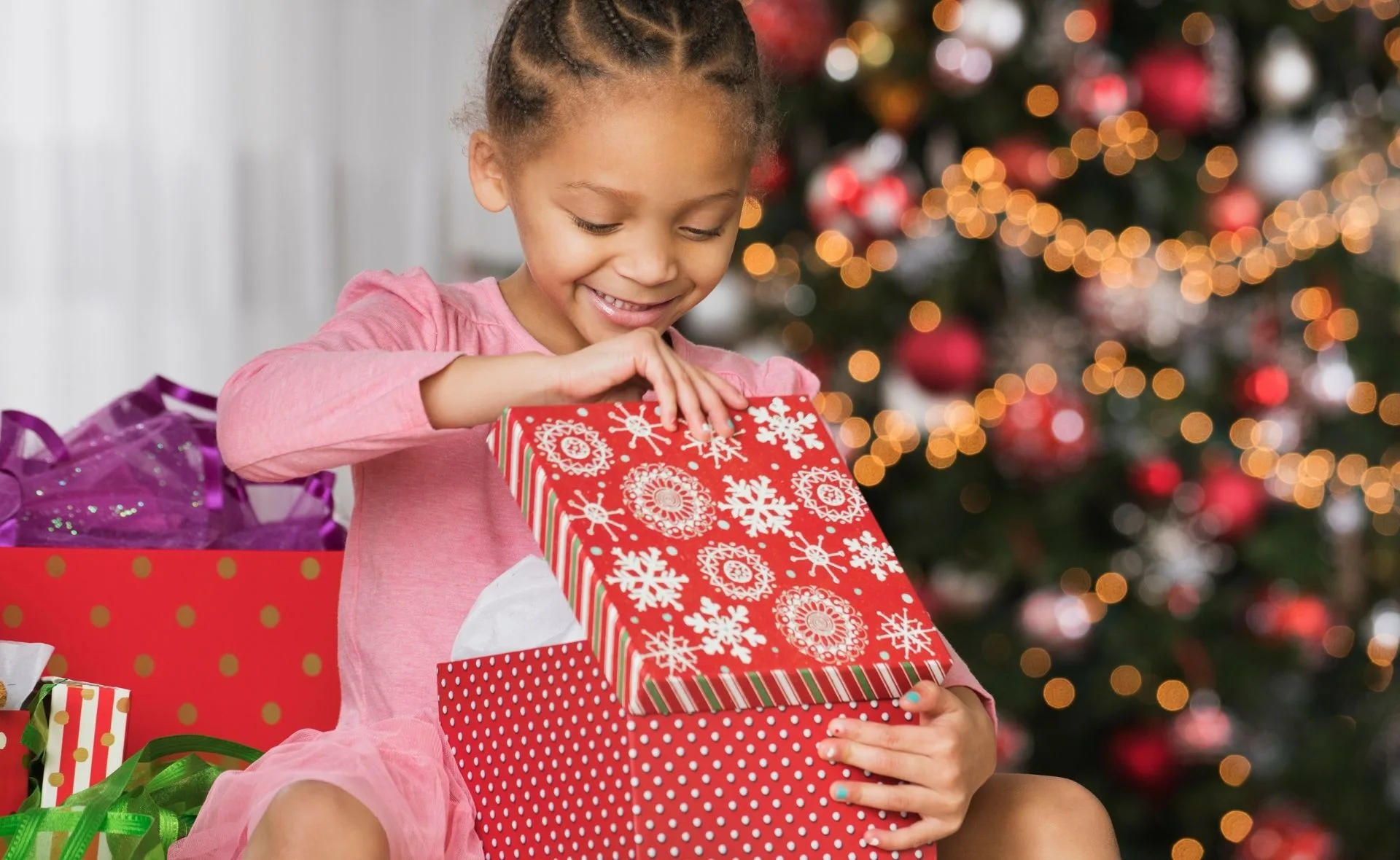 A child in a pink dress opens a patterned gift box by a decorated Christmas tree with lights.