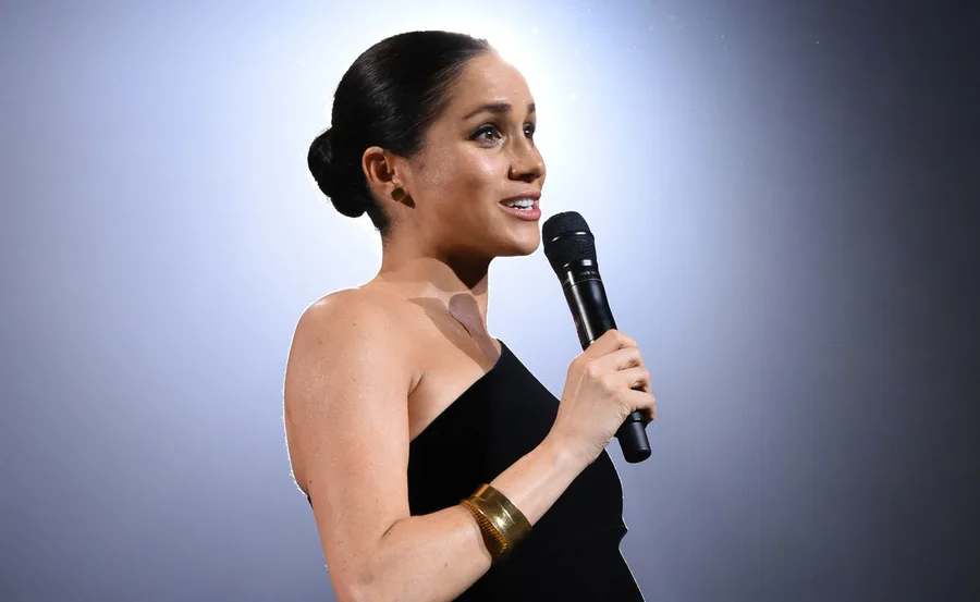 Meghan Markle holding a microphone, speaking at an event, wearing a black sleeveless dress and gold bracelet.