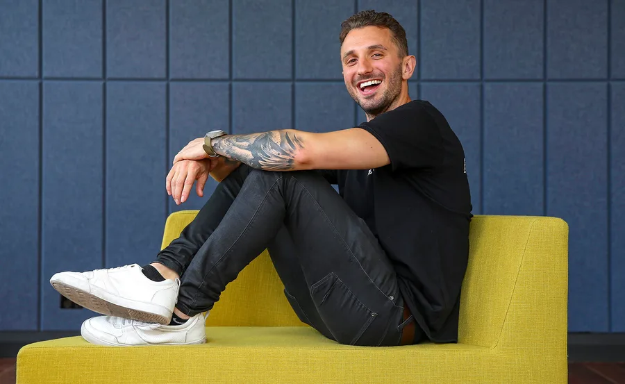 A smiling man with a tattoo on his arm, sitting casually on a yellow couch, wearing a black shirt and jeans.