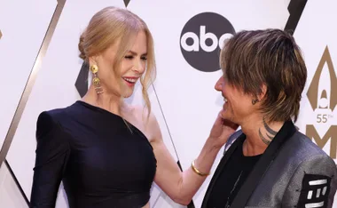 A woman with blonde hair and elegant black dress smiles at a man with brown hair and patterned suit at an event.