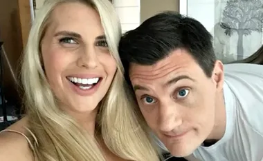 Two people smiling, a blonde woman and a dark-haired man, close together indoors.