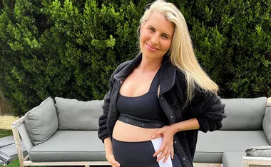 Pregnant woman in activewear smiling, standing outside by a sofa with greenery in the background.