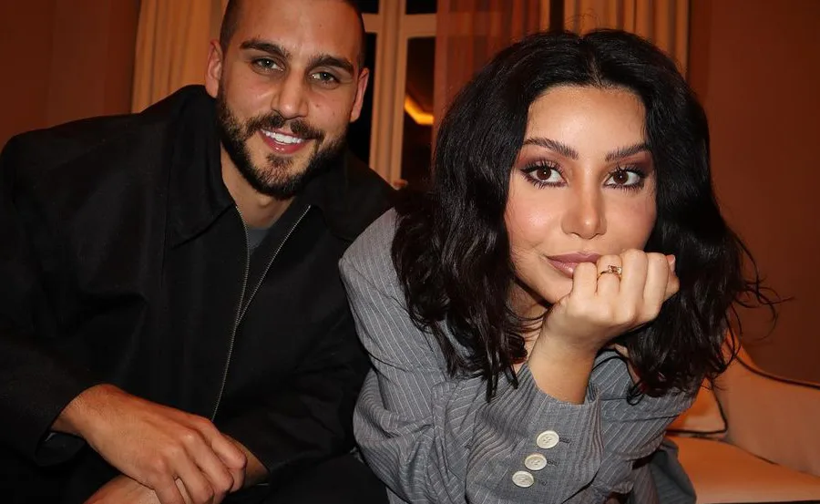 A couple smiling, sitting closely on a sofa; the woman shows her engagement ring.
