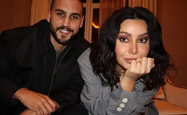 A couple smiling, sitting closely on a sofa; the woman shows her engagement ring.