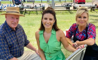 Three people sit on outdoor chairs, smiling, with a grassy background and parked cars.