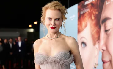 Actor in a strapless gown at an event, with a movie poster featuring a red-haired character in the background.