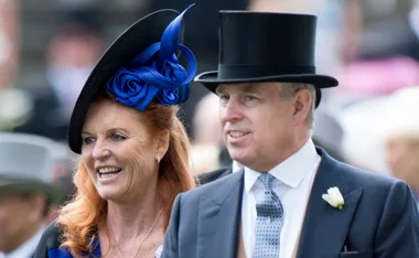 A man and woman smiling at a formal event, both wearing hats, with the woman in a decorative black hat and the man in a top hat.