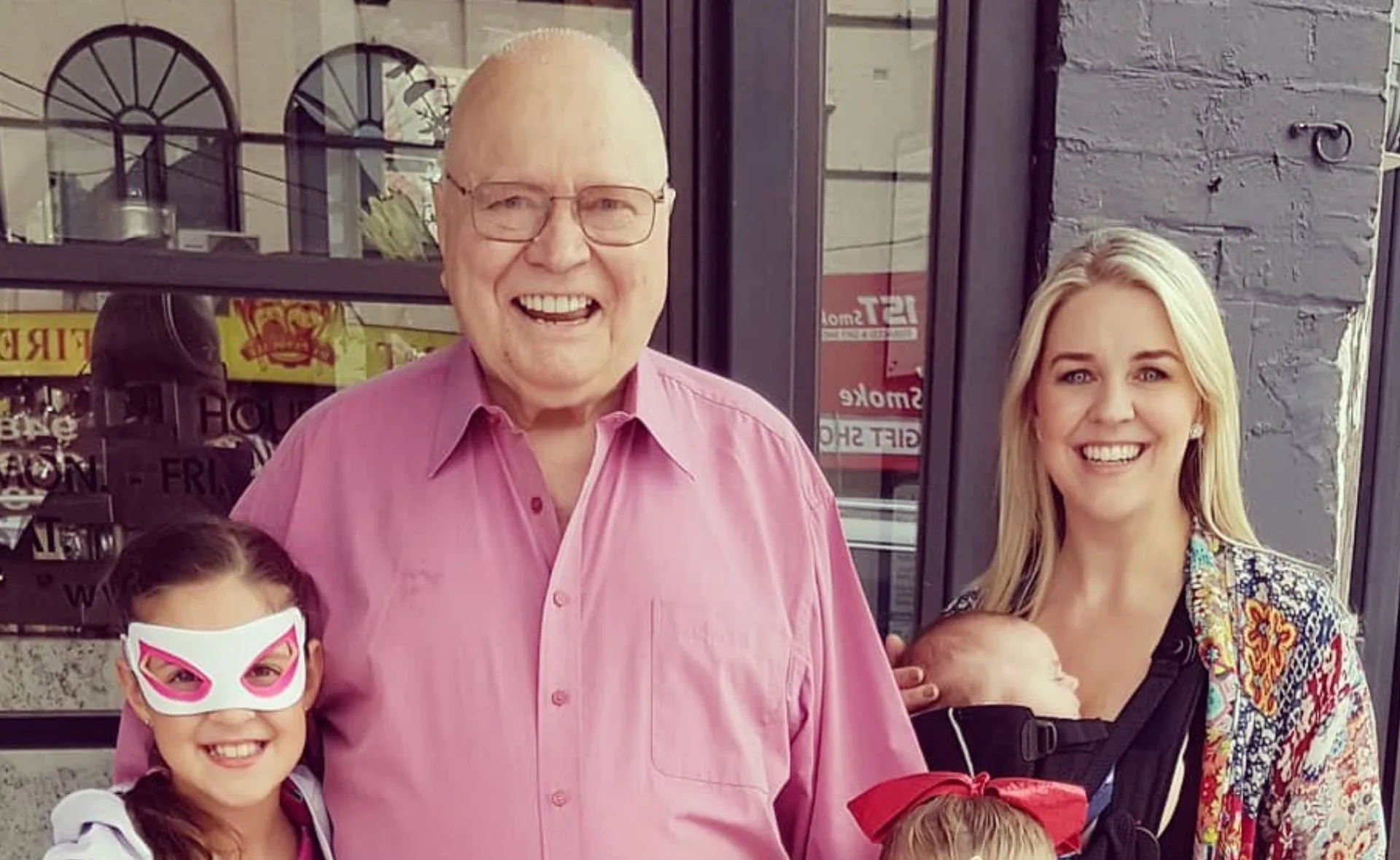 Man in pink shirt smiling with three children, one wearing a mask, another held by woman, outside a building.