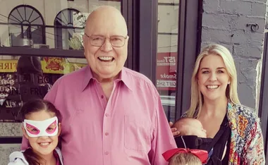 Man in pink shirt smiling with three children, one wearing a mask, another held by woman, outside a building.