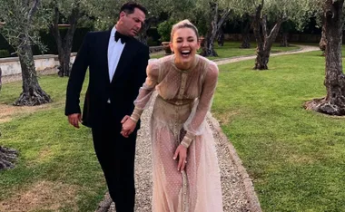 Couple holding hands, walking on a garden path; woman laughing joyfully in a sheer dress, man in a black suit.