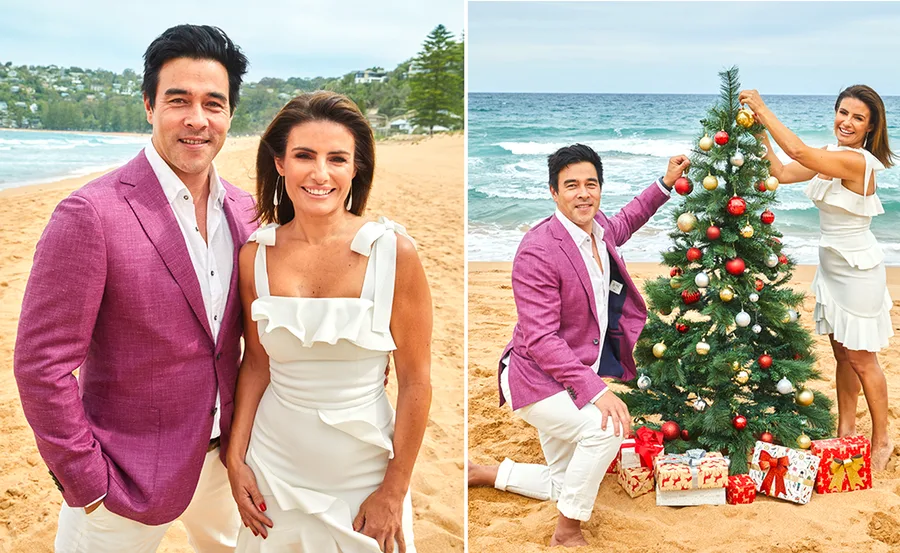 Actors on a beach, one in pink jacket, one in white dress, decorating a Christmas tree with gifts around.