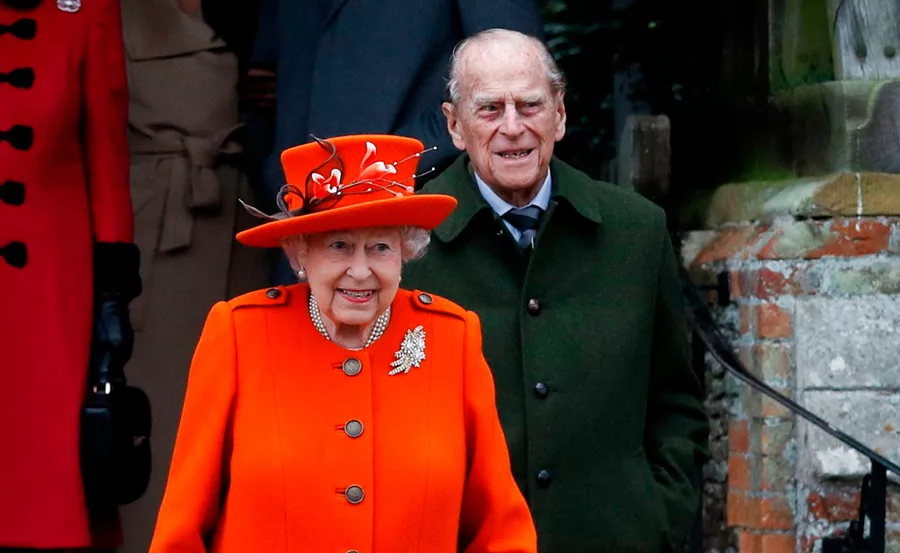 Queen Elizabeth II in an orange outfit and hat, with an older man in a green coat, outside a stone building.