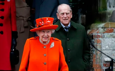 Queen Elizabeth II in an orange outfit and hat, with an older man in a green coat, outside a stone building.