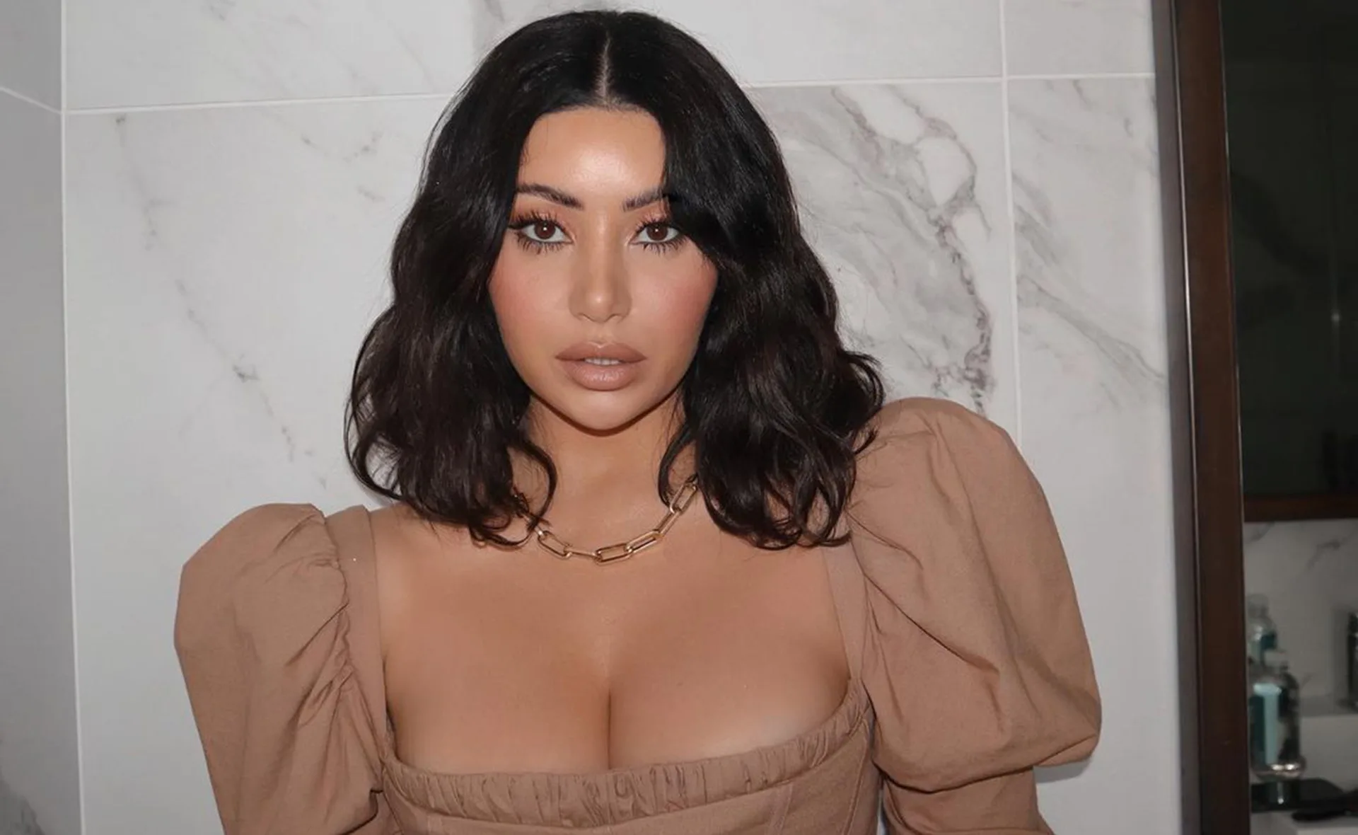 A woman with dark hair wearing a brown top and a gold necklace stands against a marbled background.