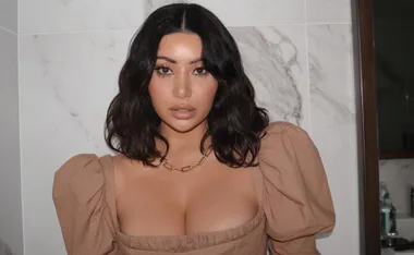 A woman with dark hair wearing a brown top and a gold necklace stands against a marbled background.