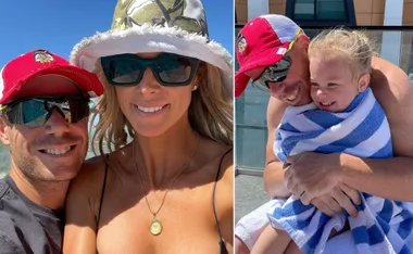 Man and woman in sunglasses smile outdoors; man hugs child wrapped in a blue-striped towel, smiling.