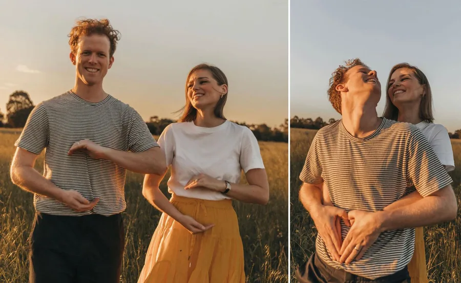 A couple standing in a field, woman in white top and yellow skirt, both forming heart shapes with hands on man's stomach.