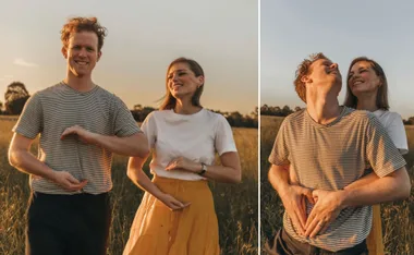 A couple standing in a field, woman in white top and yellow skirt, both forming heart shapes with hands on man's stomach.