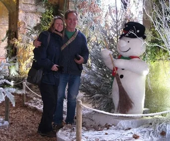 A couple poses with a snowman statue, surrounded by festive, wintry decorations and frosted trees.