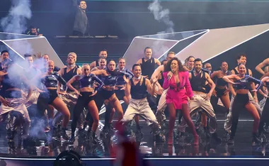 Performer in a pink outfit sings on stage with energetic dancers and a DJ, surrounded by dramatic lighting and smoke effects.