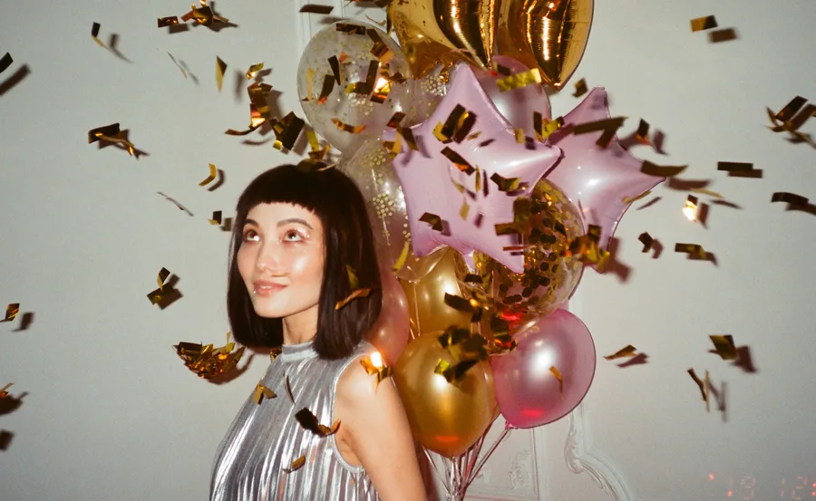 Woman in a silver dress smiling among falling confetti and colorful balloons at a celebration.