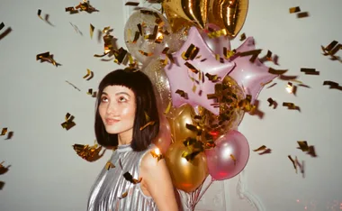 Woman in a silver dress smiling among falling confetti and colorful balloons at a celebration.