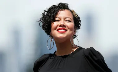 A person with curly hair and large earrings smiles brightly at the camera against a blurred background.