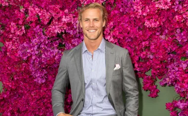 Man in a grey suit smiling in front of vibrant magenta bougainvillea blossoms.