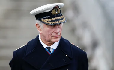A person in a naval uniform and cap, looking thoughtful, with a blurred background.