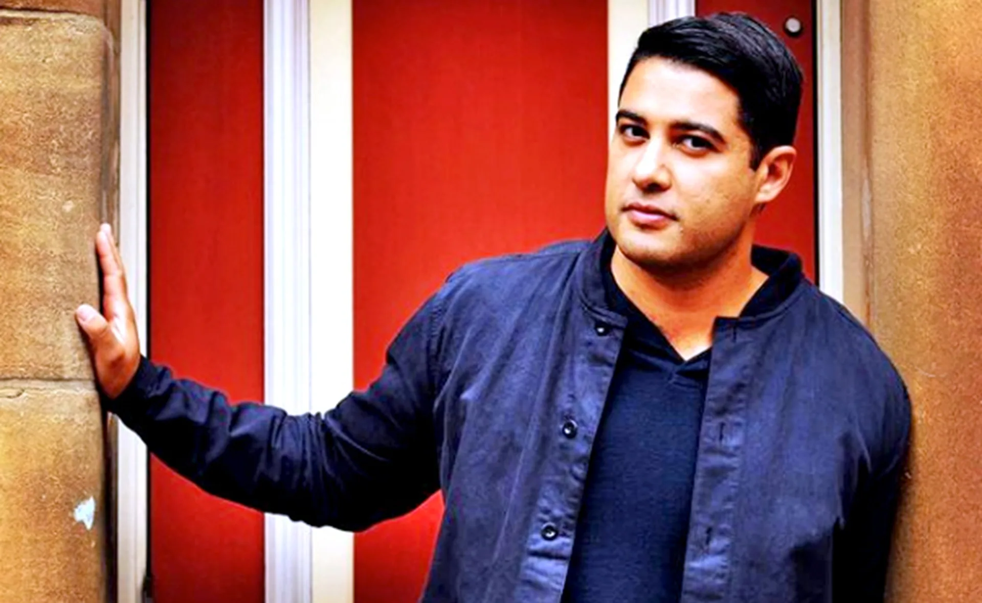 A person standing with one hand on a wall, wearing a dark jacket, in front of a red and white background.