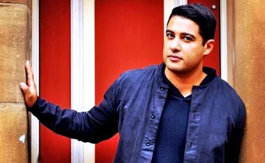 A person standing with one hand on a wall, wearing a dark jacket, in front of a red and white background.