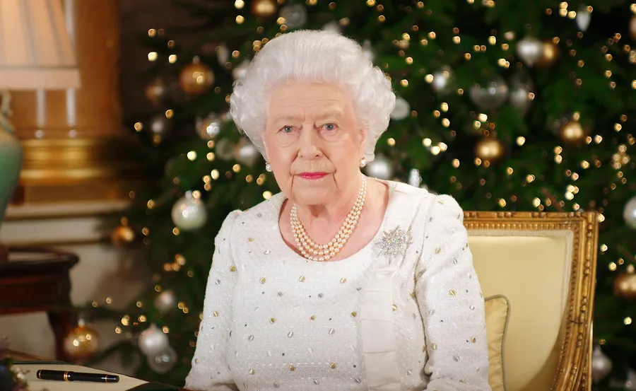 An elderly woman with white hair wearing a pearl necklace sits in front of a decorated Christmas tree.