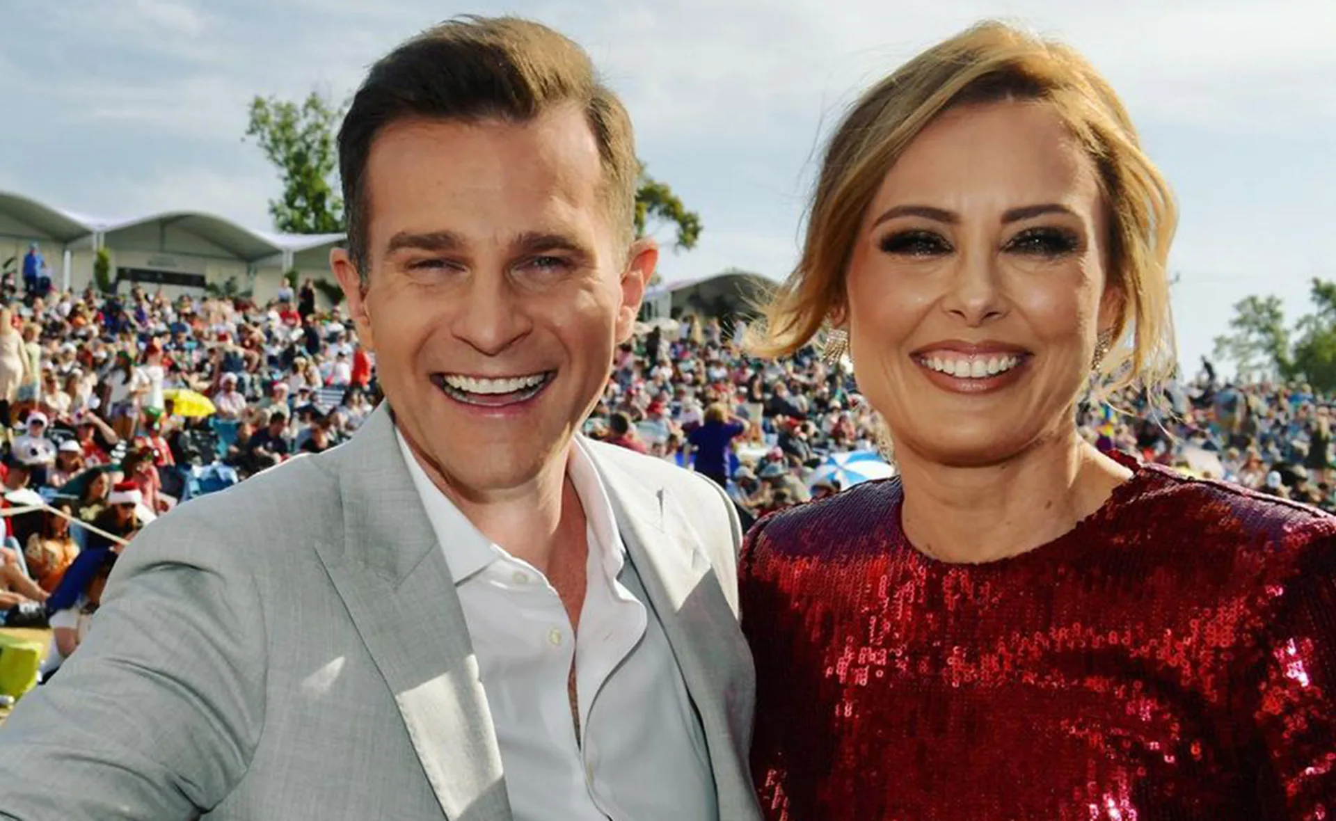 Two people smiling at an outdoor event with a large crowd in the background.