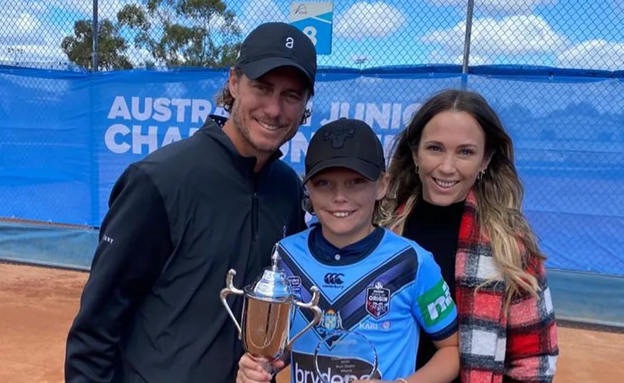 A family poses proudly as a child holds a trophy on a tennis court, with a championship banner in the background.