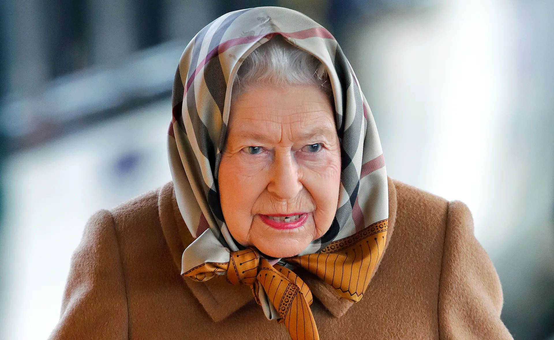 Elderly woman with a patterned headscarf and beige coat looks ahead, outdoors in natural light.