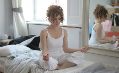 A woman in white meditating on a bed, reflected in a mirror.