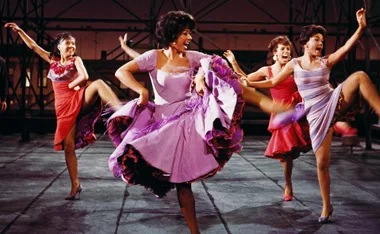 Rita Moreno as Anita dances energetically in a vibrant purple dress with fellow dancers on a rooftop in "West Side Story."