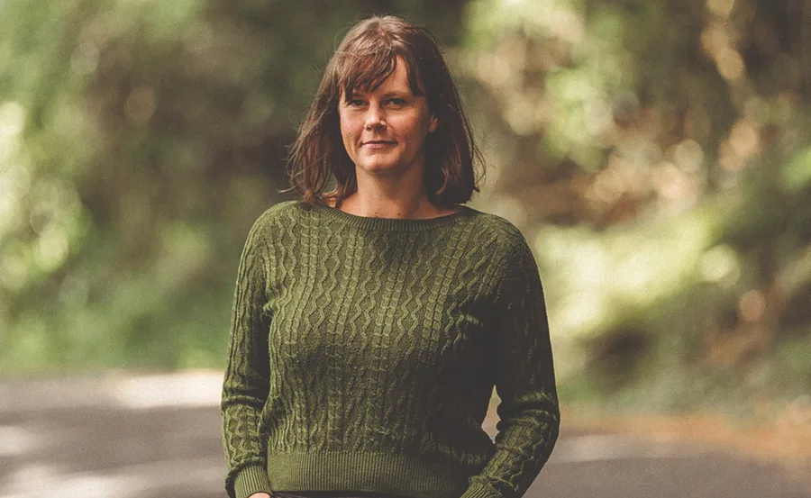 A person wearing a green sweater stands on a path with blurred trees in the background.