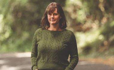A person wearing a green sweater stands on a path with blurred trees in the background.