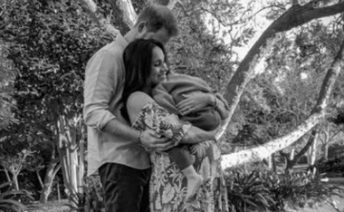 A couple stand outdoors, embracing while holding a baby wrapped in a blanket, surrounded by trees.