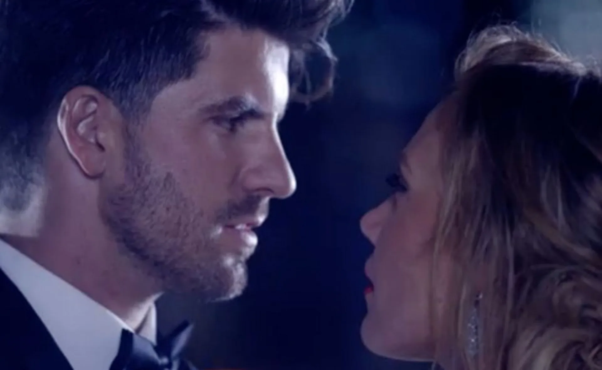 A man and woman facing each other closely in an intense moment, dressed formally, under dramatic lighting.