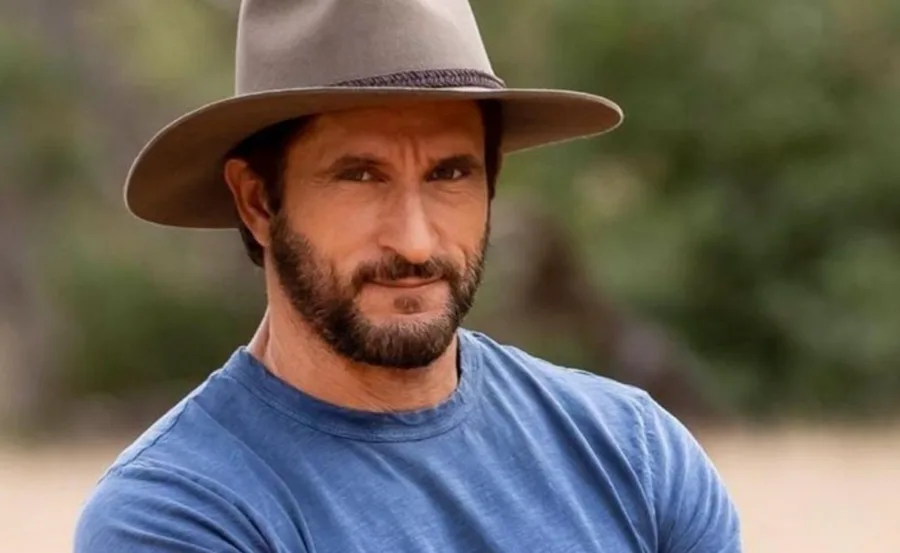 Outdoor scene with a man wearing a hat and blue shirt, looking towards the camera with a neutral expression.