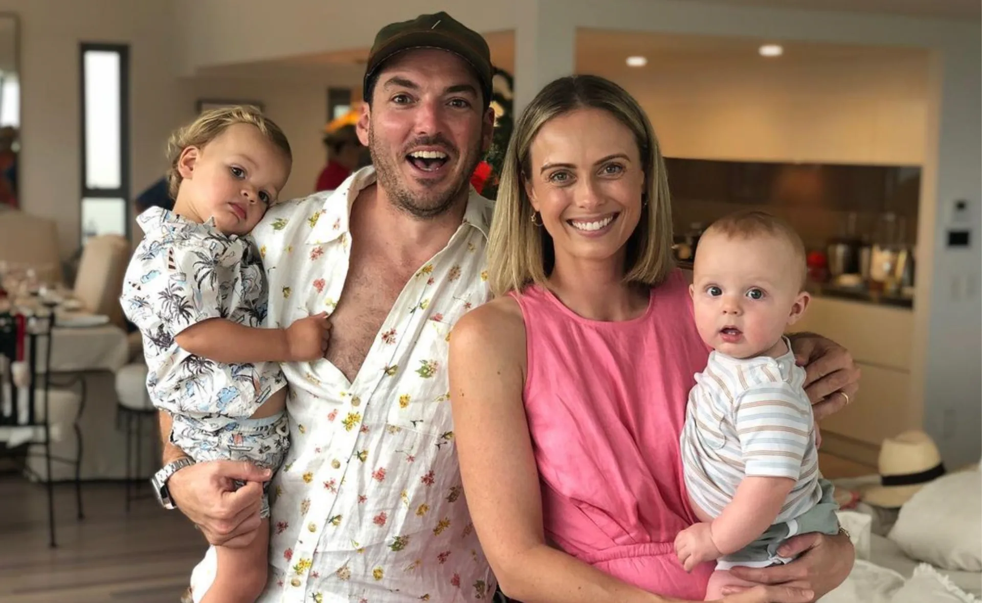 A smiling family of four in casual clothing indoors, holding their two young children.